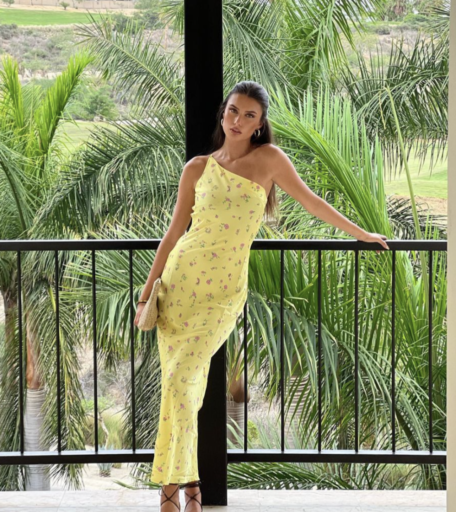 A charming one-shoulder dress adorned with a delicate floral print, perfect for a sunny day by the beach. The light yellow fabric complements the tropical surroundings, making it ideal for a relaxed yet stylish beach outing. Paired with simple sandals and a chic clutch, this outfit is the epitome of a summer dress designed for tropical beach adventures.